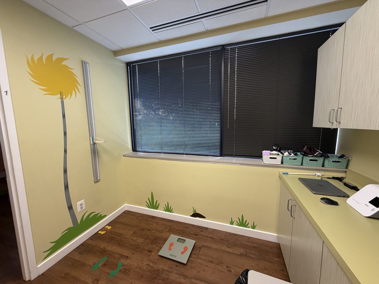 Sunny yellow exam room with a wall-mounted height board, floor scale and tree mural