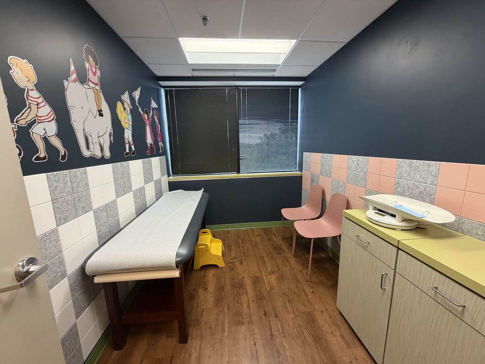Navy exam room with a storybook-parade mural, exam table and infant scale