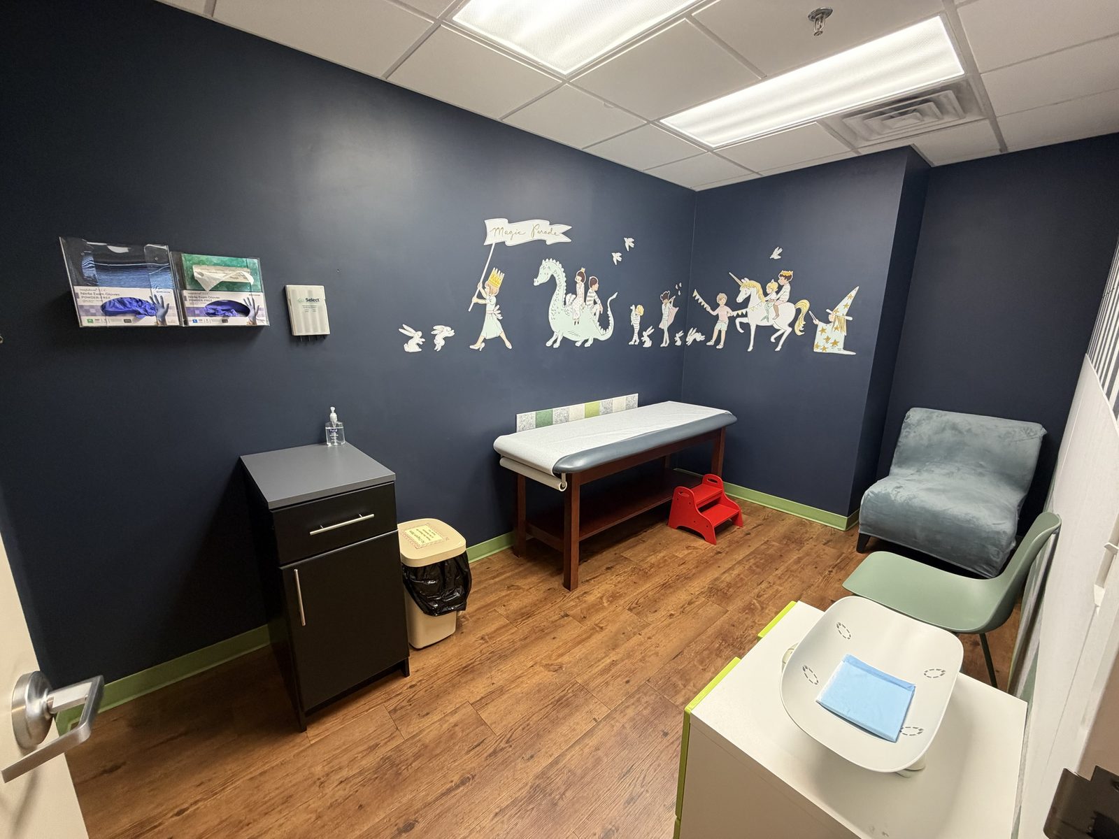 Navy exam room with a fairy-tale parade mural, a comfy armchair and an infant scale